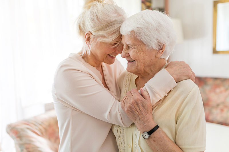 Two older women in white hugging one another in a white walled living room.